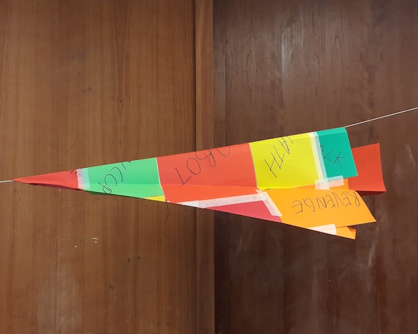 Colorful handmade paper rocket with handwritten labels, suspended on a string against a wooden background